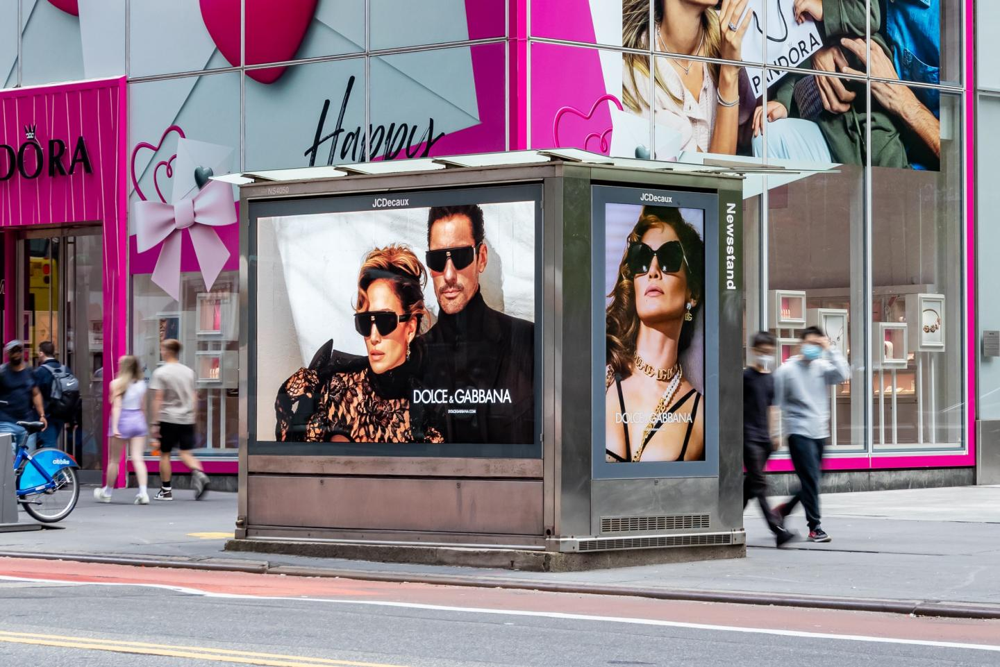 Freestanding JCDecaux newsstand billboard on a city sidewalk displays Dolce and Gabbana fashion ads featuring models in black clothing and oversized sunglasses. The kiosk stands in front of a bright pink Pandora storefront with gift box graphics while pedestrians walk past in motion blur in Times Square..