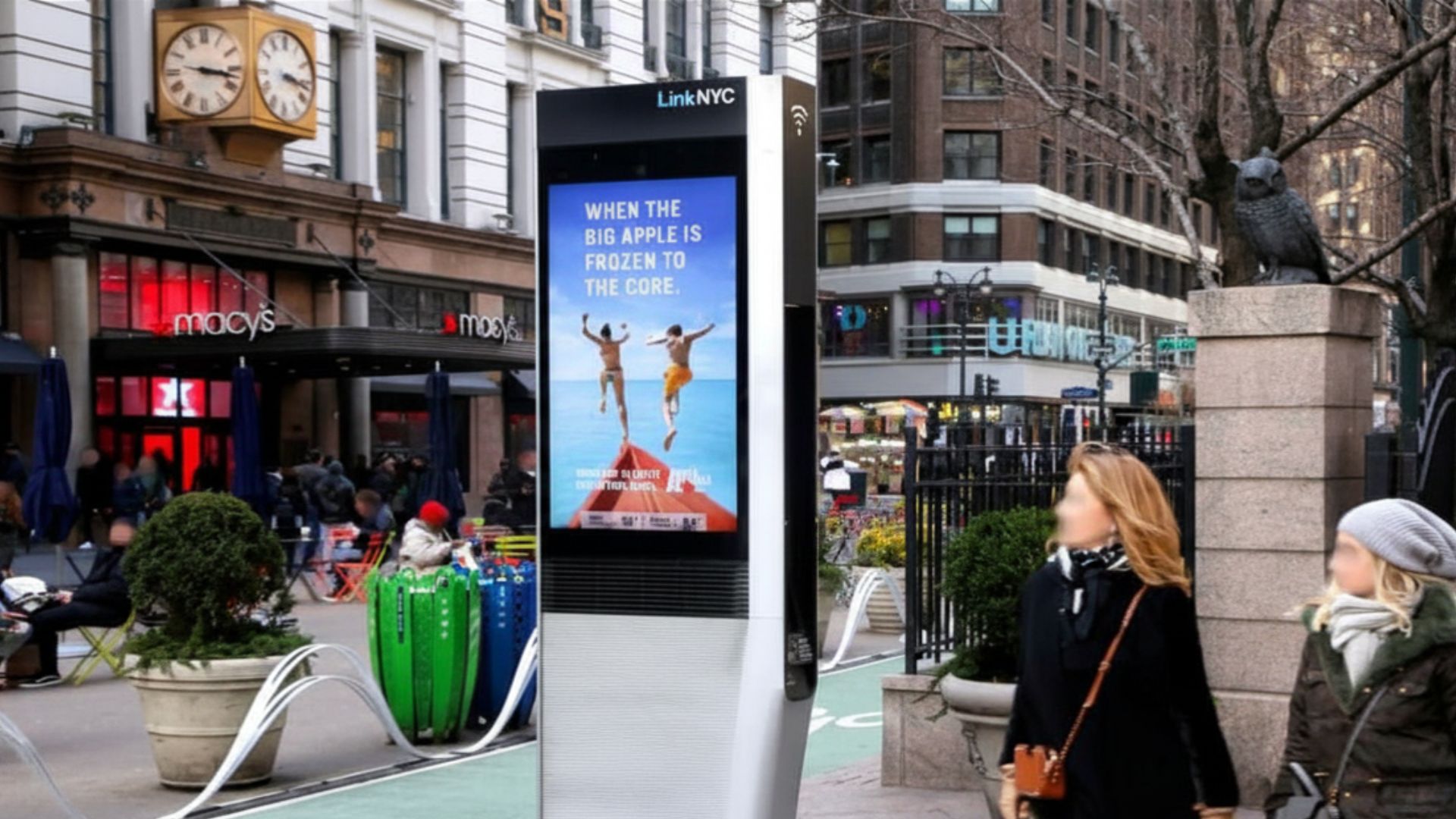 LinkNYC digital kiosk on a city sidewalk displays a winter travel ad that reads "WHEN THE BIG APPLE IS FROZEN TO THE CORE." Two women walk past the screen near Macys, planted medians, and a stone pedestal with a bird statue