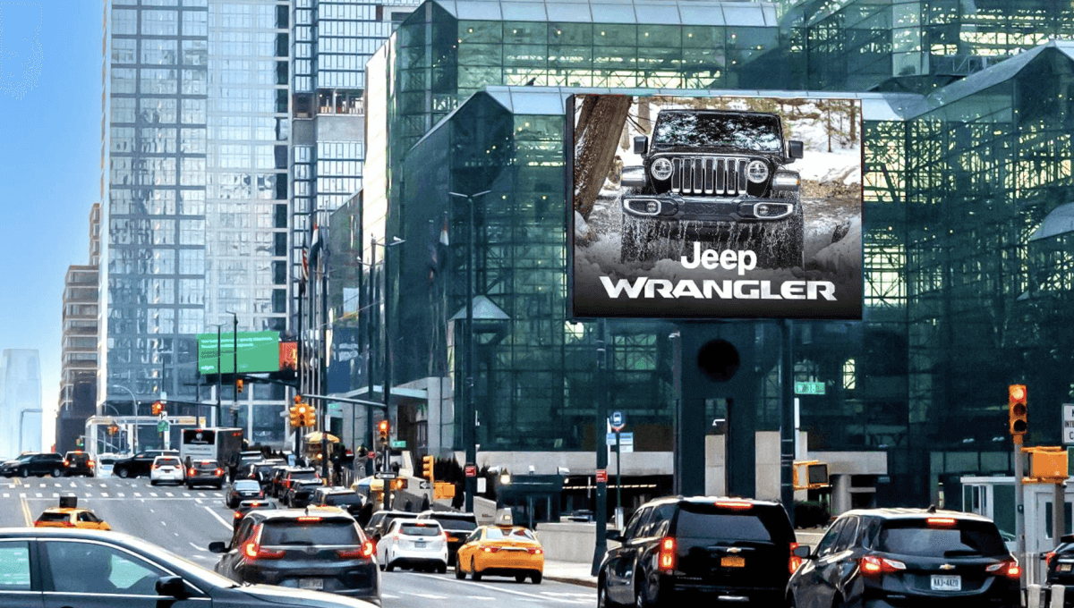 Large roadside billboard above a busy city avenue displays a Jeep ad with a front view of a Wrangler driving through snow and the text "Jeep WRANGLER." Cars and yellow taxis move below between glass office towers and a modern atrium building.