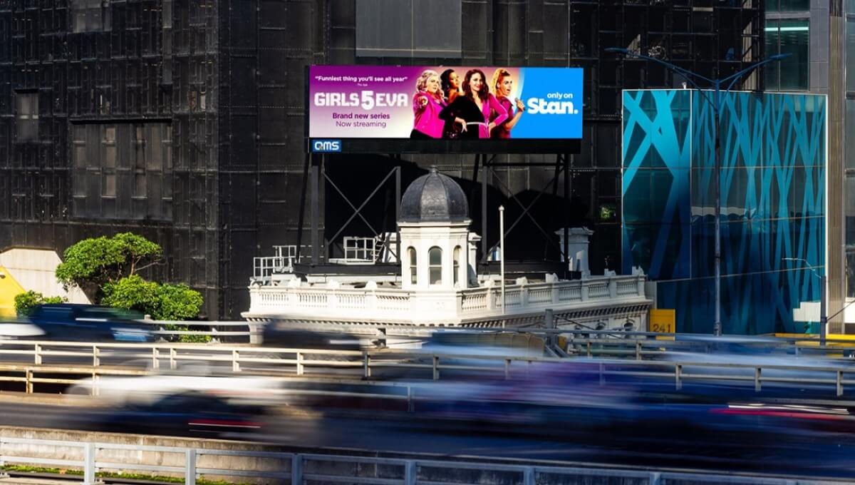 Elevated roadside billboard above a multilane roadway displays a bright pink and blue ad for "GIRLS5EVA" with four women and the text "Brand new series Now streaming" and "only on Stan." Motion blurred traffic passes below while the billboard rises behind a small white domed historic building and beside dark glass towers.