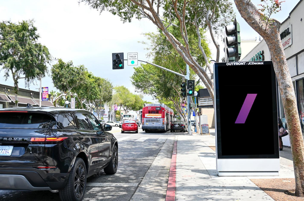 Street side digital billboard on a sunny tree lined avenue displays a minimalist ad with a single purple diagonal bar on a black background. Cars, a city bus, traffic lights, storefronts, and broad sidewalks frame the clean urban streetscape.