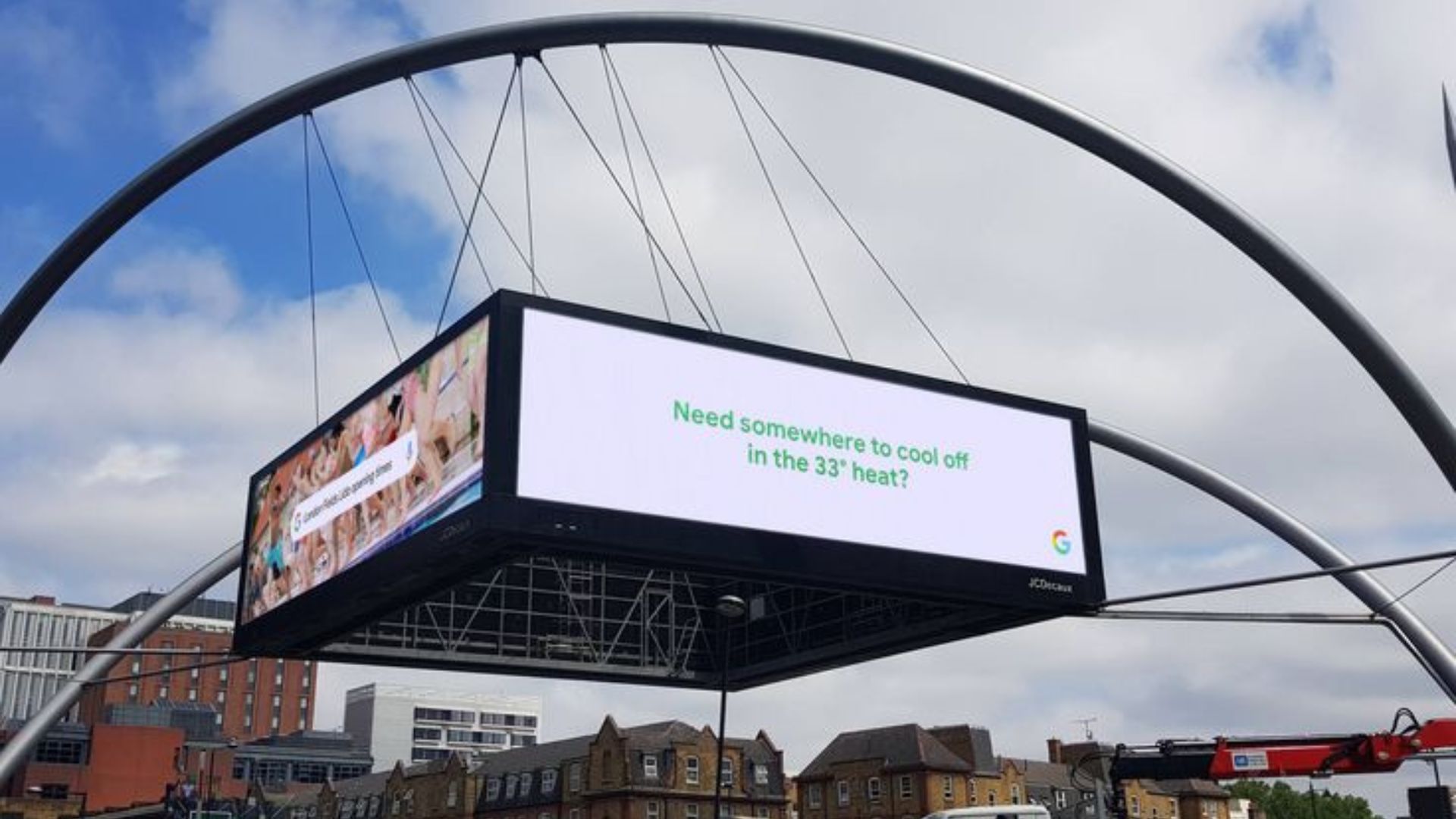 Large suspended digital billboard hangs beneath sweeping metal arches and displays a Google ad that reads "Need somewhere to cool off in the 33° heat?" against a white background. The screen floats above low city buildings under a cloudy sky with patches of blue.