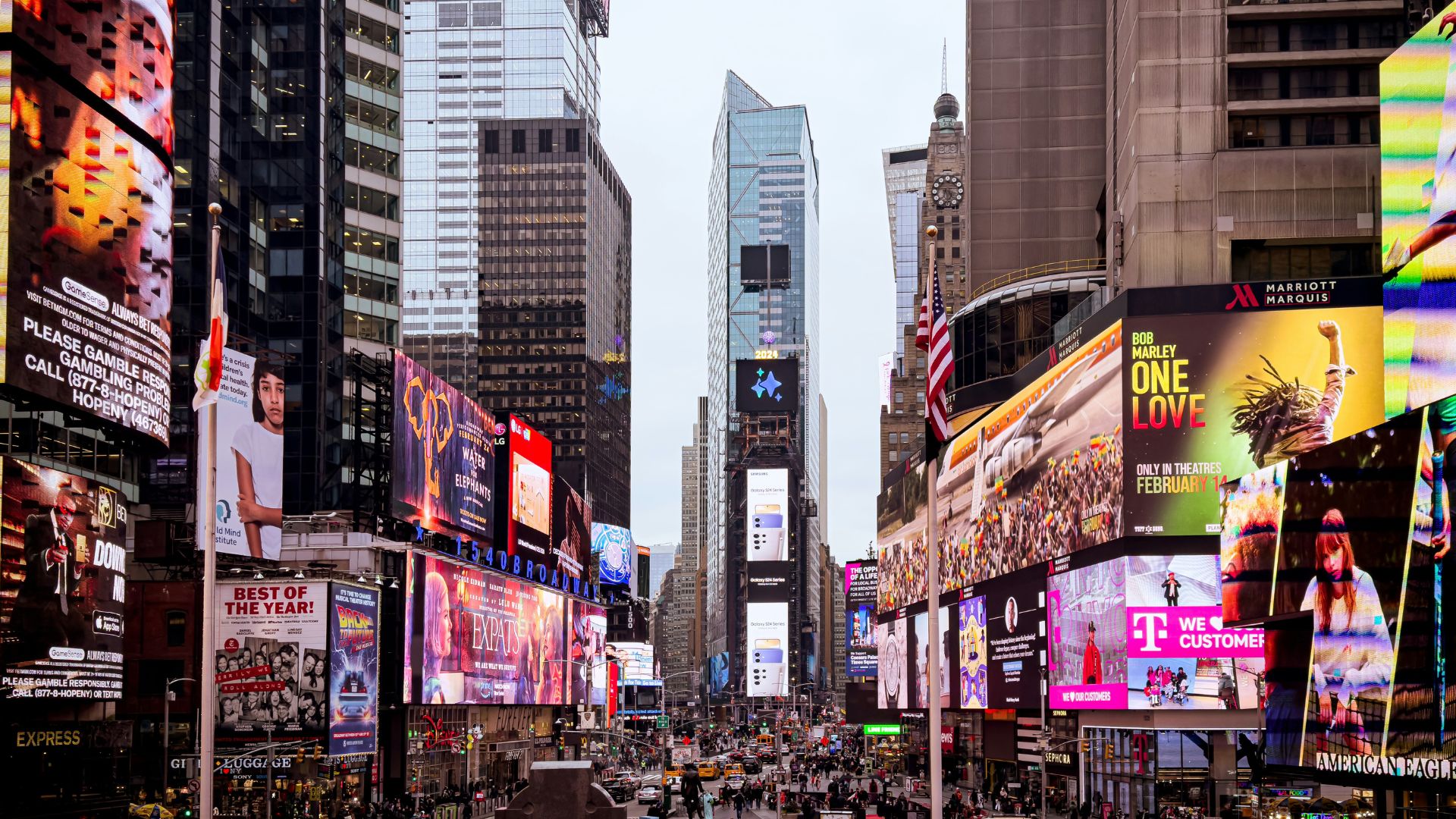 Wide view of Times Square packed with towering digital billboards, traffic, and crowds moving through the center of Manhattan. Bright ads for movies, phones, fashion, theater, and brands cover both sides of the street, including a large "Bob Marley One Love" billboard and stacked screens rising into the distance.
