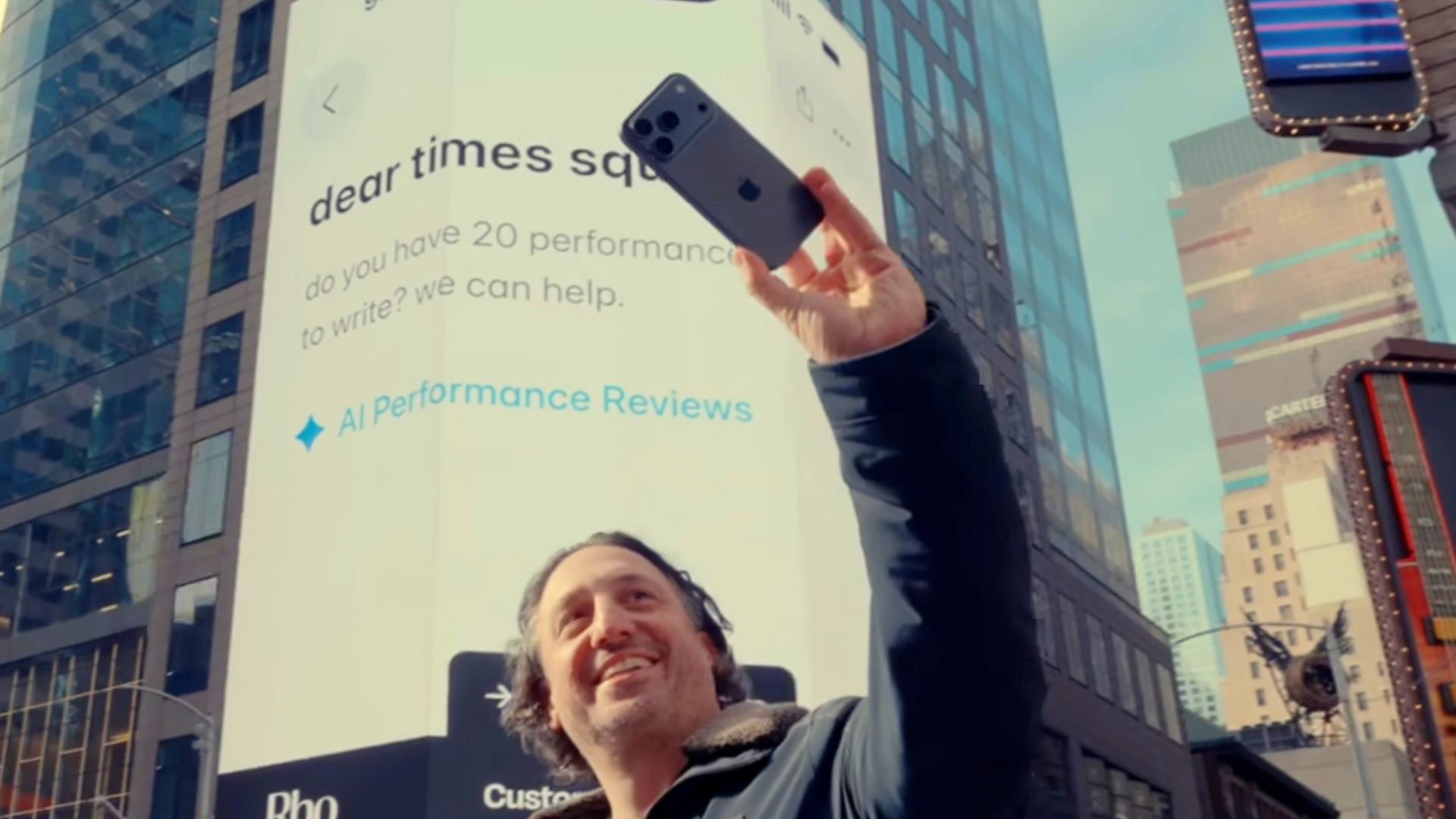 Person in Times Square holds up a smartphone for a selfie in front of a giant white billboard that reads "dear times square do you have 20 performance reviews to write? we can help." and "AI Performance Reviews." Tall glass buildings and surrounding signs frame the bright display.