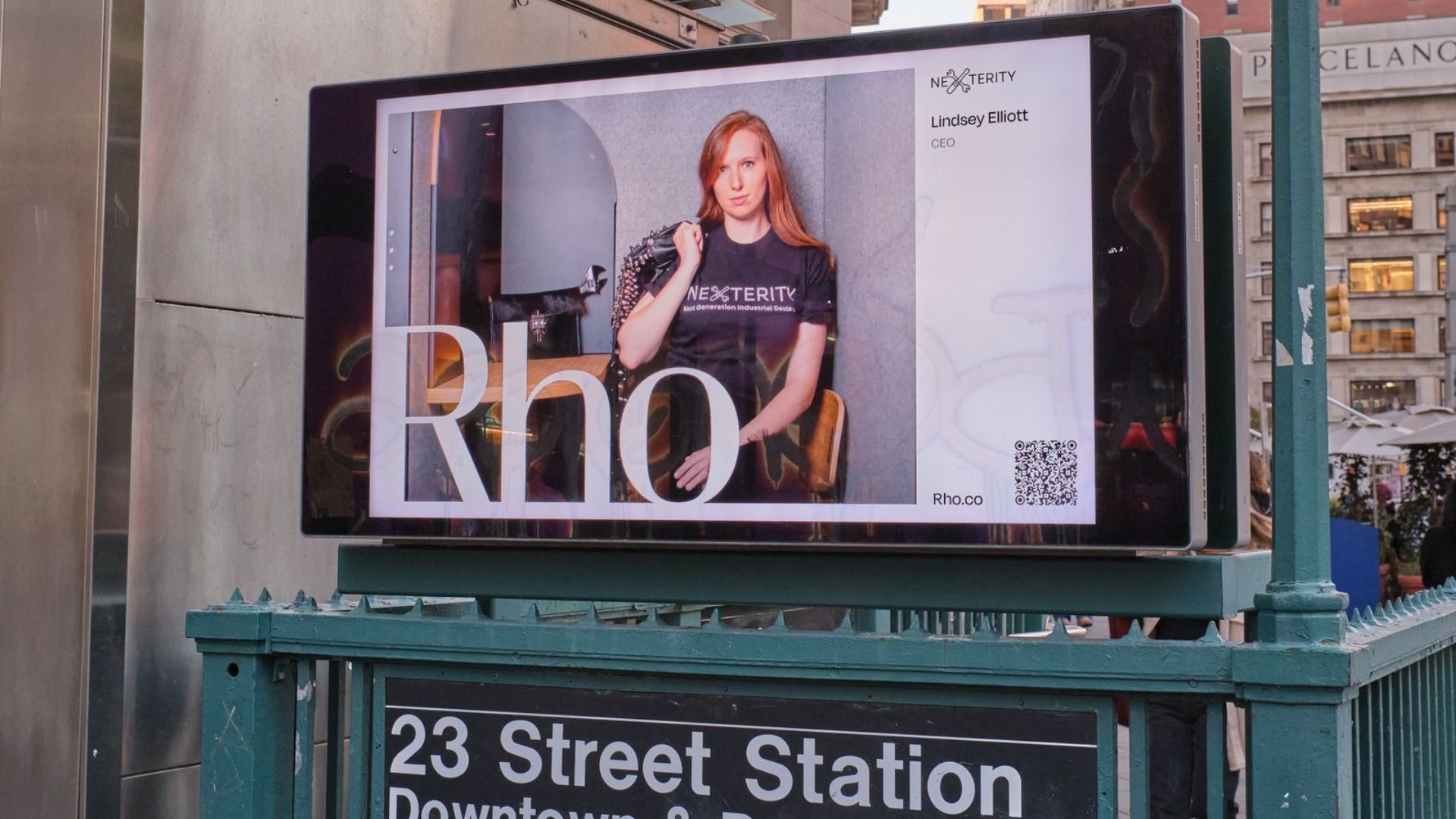 Digital billboard above the "23 Street Station Downtown and Brooklyn" subway entrance shows a portrait of a red haired woman seated in a chair wearing a "NEXTERITY" shirt. The ad includes the text "Lindsey Elliott" "CEO" "Rho" and "Rho.co" with a QR code on the right.