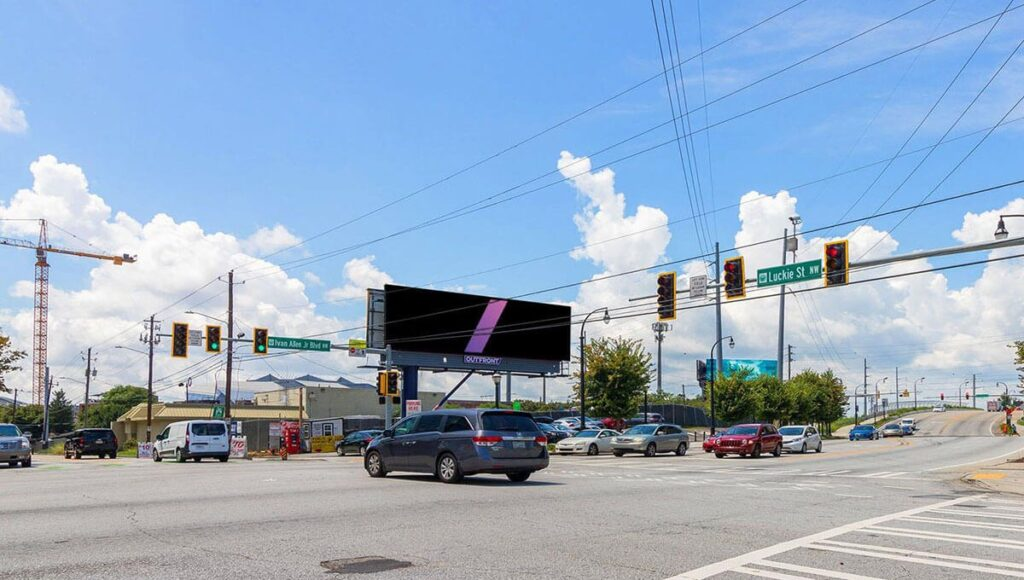Roadside digital billboard at a busy intersection displays a simple black screen with a single diagonal purple bar. Cars wait and turn below the sign beneath traffic lights and street signs for "Luckie St NW" and "Ivan Allen Jr Blvd" under a bright blue sky with large white clouds.