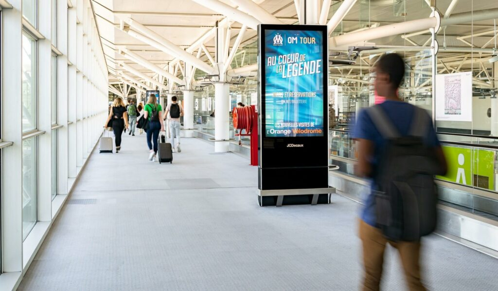 Digital billboard stands in a bright airport or transit corridor and displays an Olympique de Marseille tour ad that reads "OM TOUR" and "AU COEUR DE LA LÉGENDE." Travelers with rolling luggage walk through the glass walled passage while white beams and metal structure frame the interior.