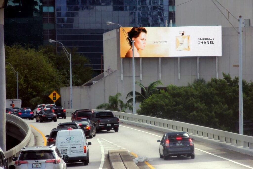 Highway billboard above a curving city road displays a Gabrielle Chanel fragrance ad with a woman's profile, a perfume bottle, and the text "The New Fragrance" and "Gabrielle Chanel." Cars move along the elevated roadway below with palm trees, greenery, and dark glass buildings surrounding the sign.