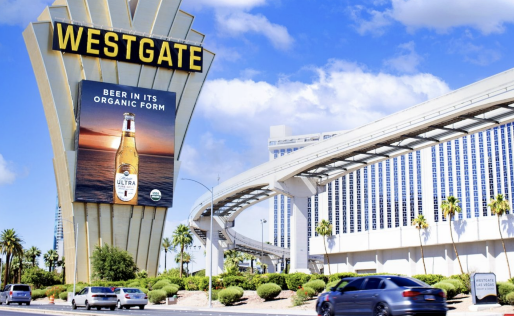 Tall Westgate tower sign against a bright blue sky displays a Michelob Ultra ad with a beer bottle over a sunset water scene and the text "BEER IN ITS ORGANIC FORM." A curved elevated monorail track sweeps past the hotel building on the right while cars and palm trees line the boulevard below.