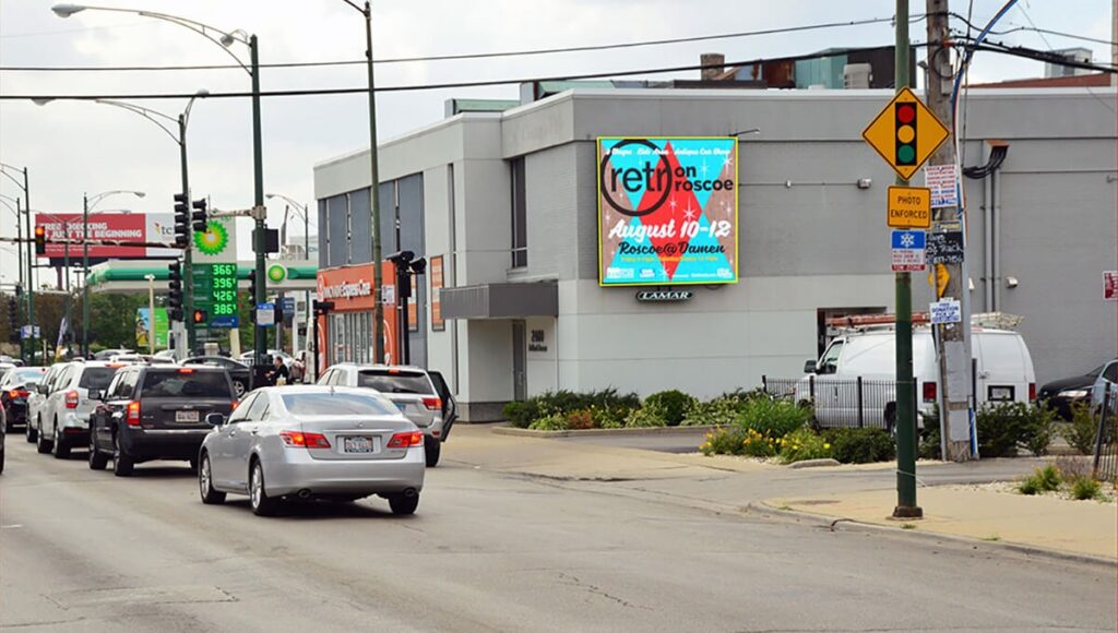 Roadside billboard mounted on the side of a low gray building displays a colorful poster for "retro on roscoe" with the dates "August 10-12" and the line "Roscoe at Damen." Cars move through the city street below while traffic lights, utility wires, and storefronts line the intersection.