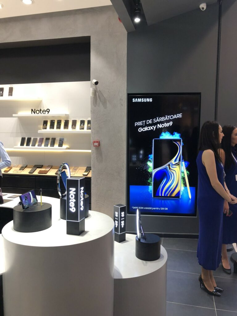 Samsung store interior with a vertical digital display advertising the Galaxy Note9 in Romanian. The screen reads "SAMSUNG" and "PREȚ DE SĂRBĂTOARE Galaxy Note9" beside a phone image, while nearby shelves labeled "Note9" and lit product stands display phones and accessories as two women in blue dresses stand to the right.