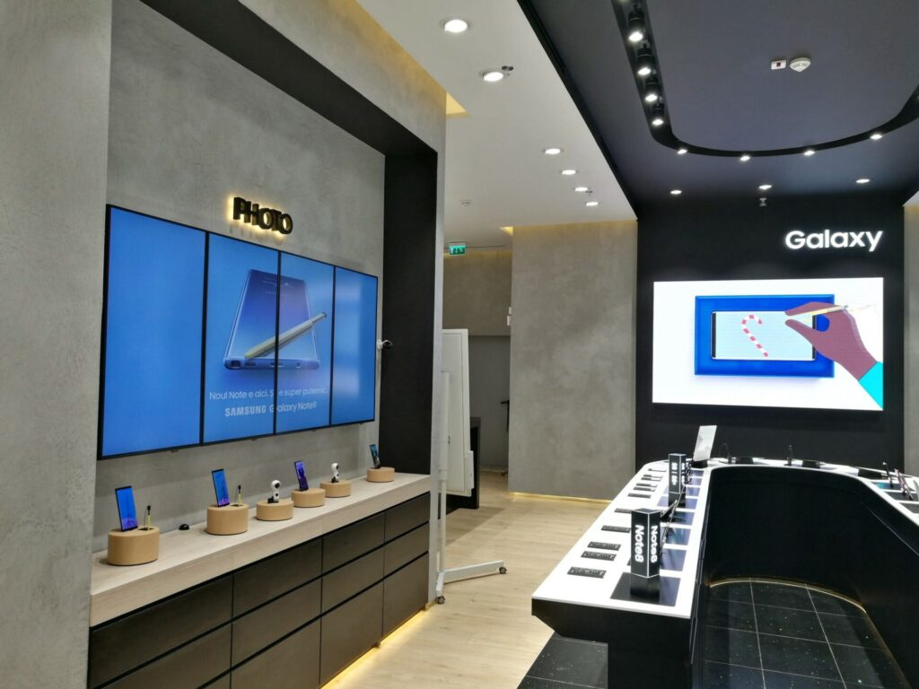 Modern Samsung store interior with illuminated displays for the Galaxy Note9 and a wall section labeled "Photo." Three large vertical screens on the left show a Note9 ad, while a bright screen at the back under the "Galaxy" sign shows a hand drawing on a phone and demo devices line the counters below.